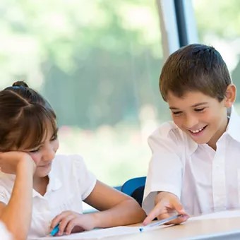A boy and girl in school uniforms sit together at a desk, smiling and interacting over their work in a bright classroom setting.