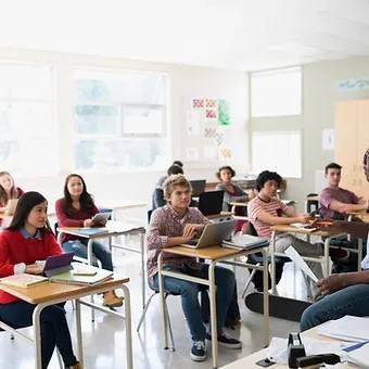 A diverse group of high school students sit at desks in a bright classroom, attentively listening to a teacher who is partially visible in the foreground.