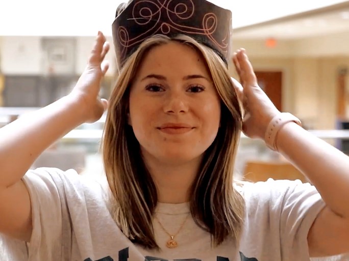 A smiling student happily places an ornate hat upon their head.