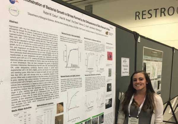 Photo: Robin Cesur stands with the poster presentation demonstrating that microbes contaminating spacecraft can grow in brines formed by the deliquescent wetting of salts by humidity alone.