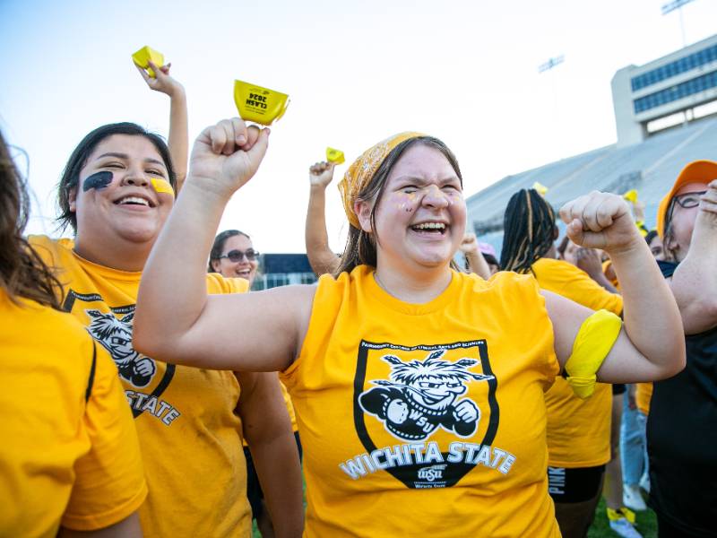 Smiling Fairmount College students in yellow Wichita State shirts cheer at Clash of the Colleges.