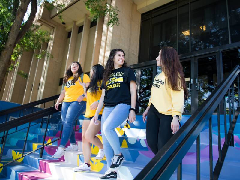 Four students in Wichita State University shirts walk down colorful outdoor steps, smiling and talking.