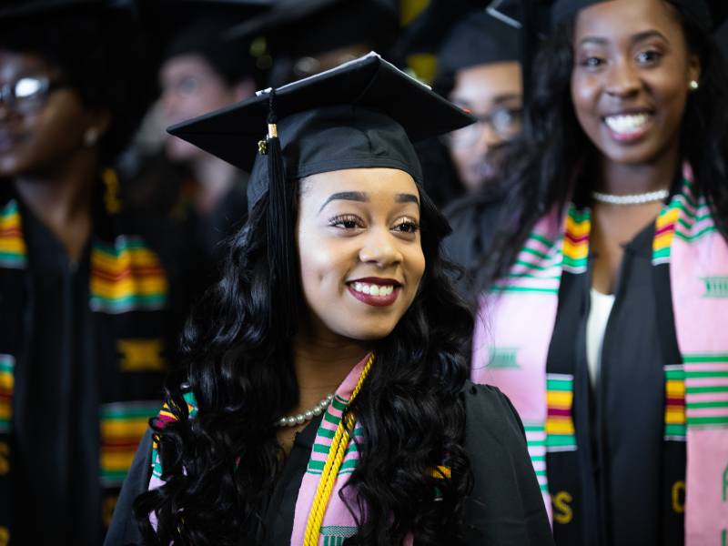 Smiling female student in a graduation cap and gown stands with classmates during a graduation ceremony.