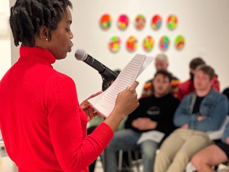 A woman wearing a red sweater reads from a paper while speaking into a microphone at an open mic event. Several seated audience members listen in the background.