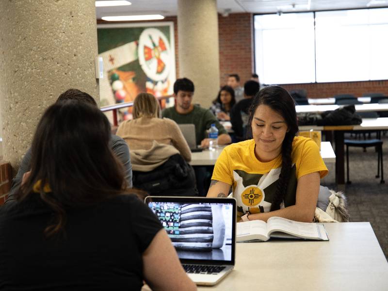 Students study together at a table in a library, with one student reading a book and another using a laptop.