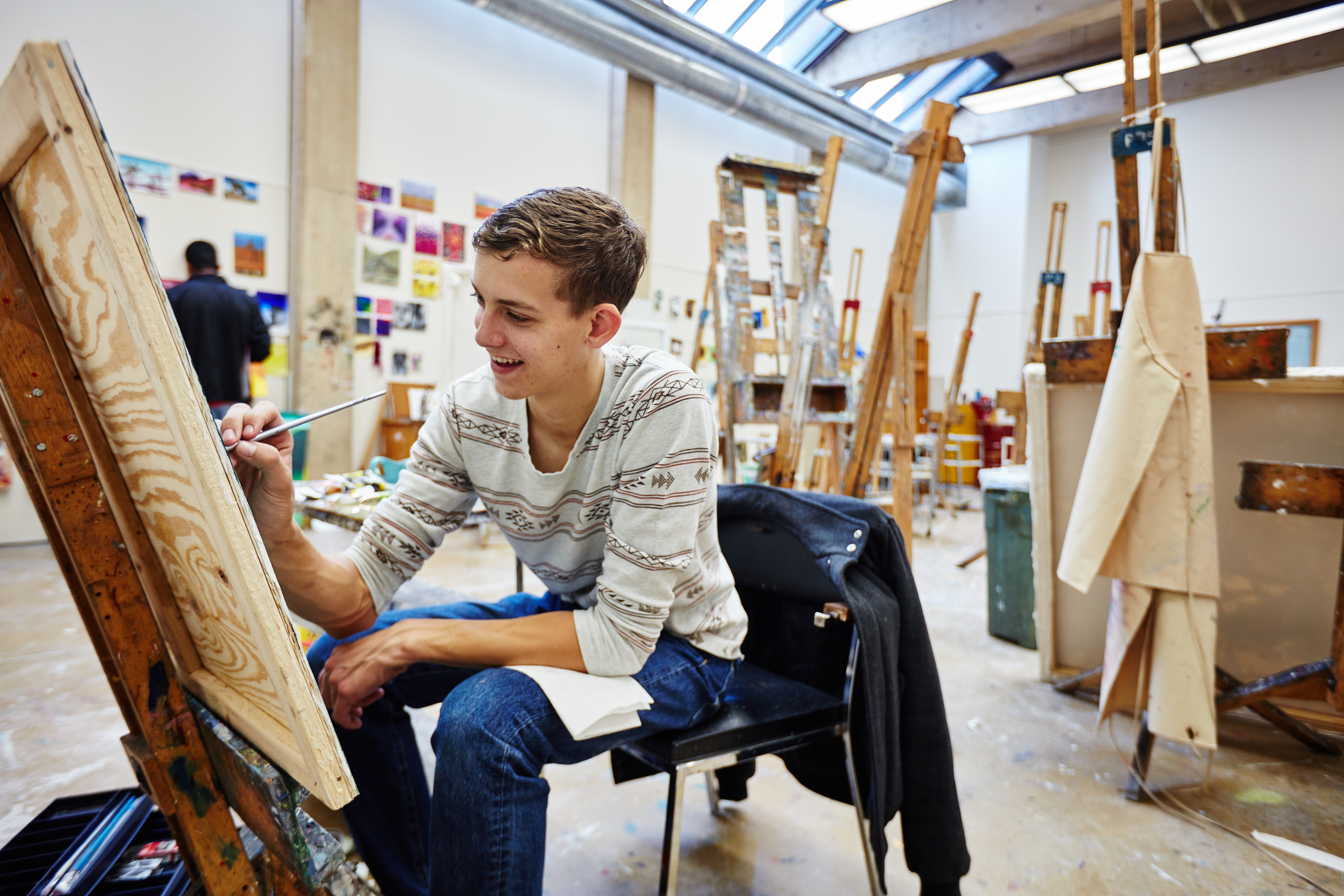 Smiling figure sitting at easel painting.