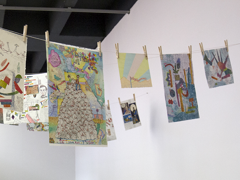 A photograph of several colorful prints displayed in the PrintSpace gallery. The prints are attached to a line with wooden clothespins.