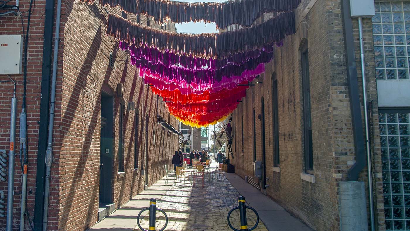 A photograph of Gallery Alley, an outdoor arts space in downtown Wichita. Several strands of vinyl fringe line the alley, creating a gradient affect. The alley's cobblestones are painted and it's full of brightly colored bistro tables and chairs. 