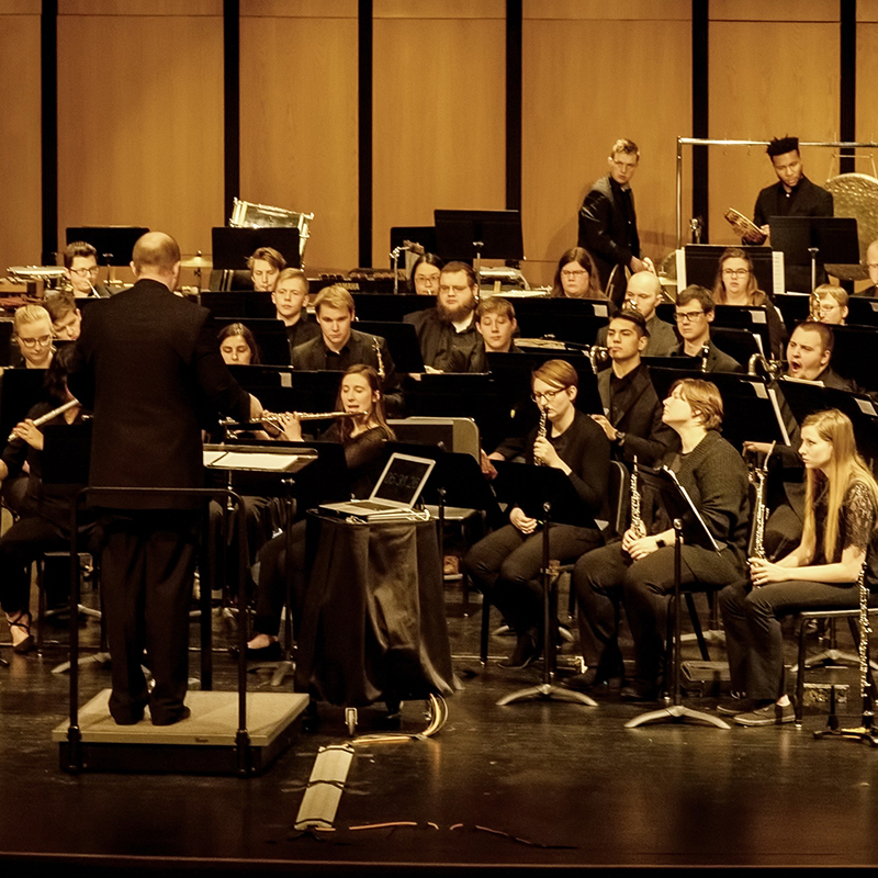 A picture of a university ensemble amidst a rehearsal or performance. The is a conductor on the left side of the photo and most of the musicians are on the right side of the picture.