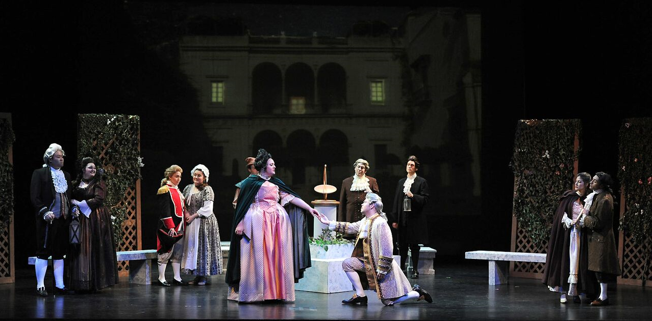 A photo of a opera performance with several performers on stage. Mostof the actors and actress are dressed in 18th century attire, with the focus of the photograph being a man and woman in the center. The man is kneeling in front of the woman, holding her hand in his hand.