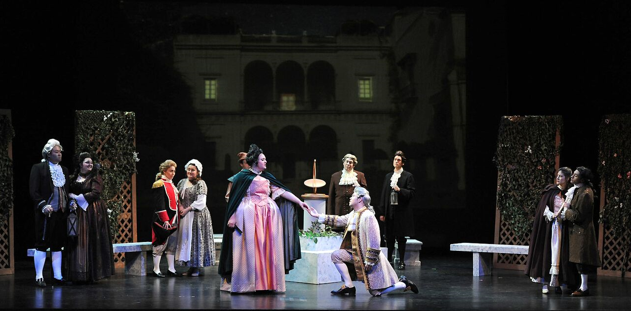 An image of Opera performers on stage during a performance. One actor is on his knees with a woman's hand in his hand while he sings to her. There are other performers in the background watching the scene unfold.