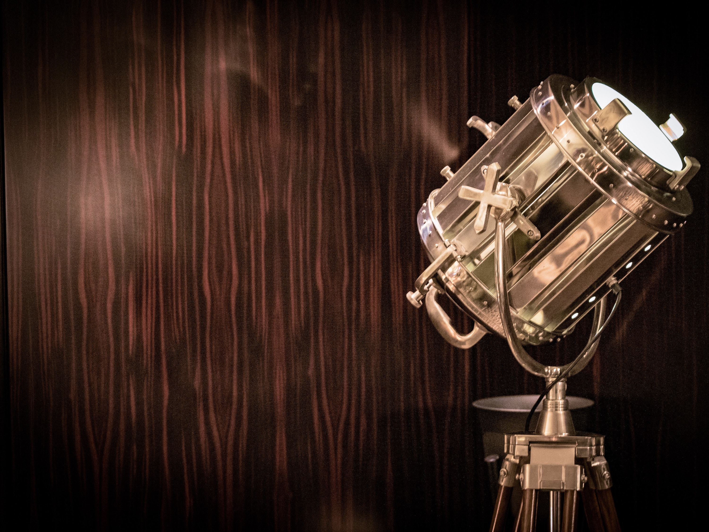Photo of a large, illuminated brass spotlight shining light toward the upper right with a dark wood panelled wall behind it. 