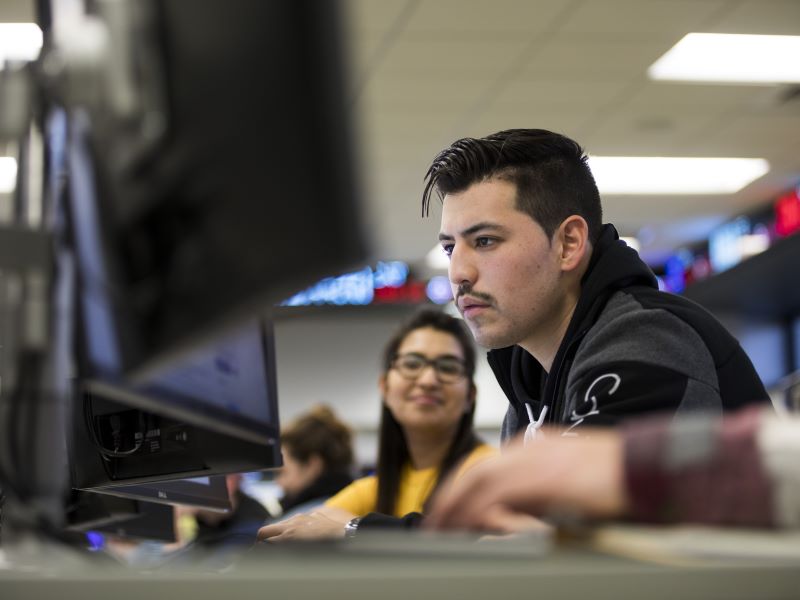 Man focused on a computer screen in a classroom, with another student smiling in the background.