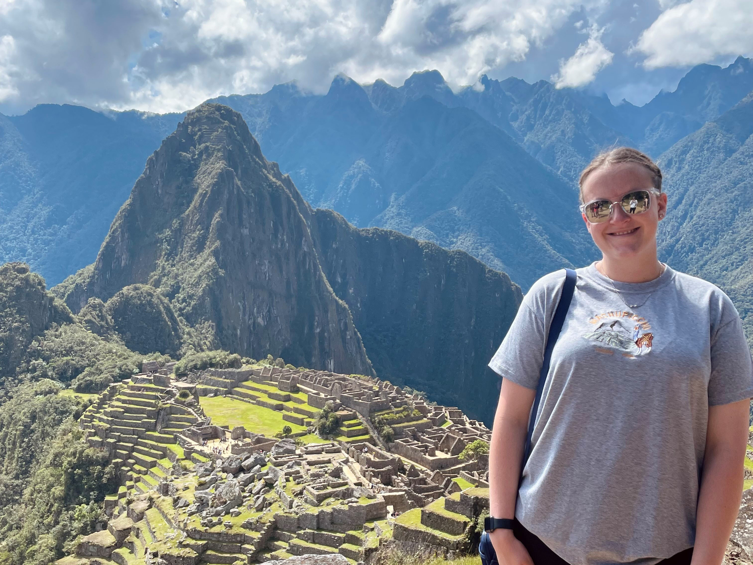 Honors student Katie Dunlop posing for a photo in Machu Picchu, Peru. Katie is wearing a gray t-shirt, black leggings, and black refelective sunglasses. Machu Picchu is featured prominently behind her.