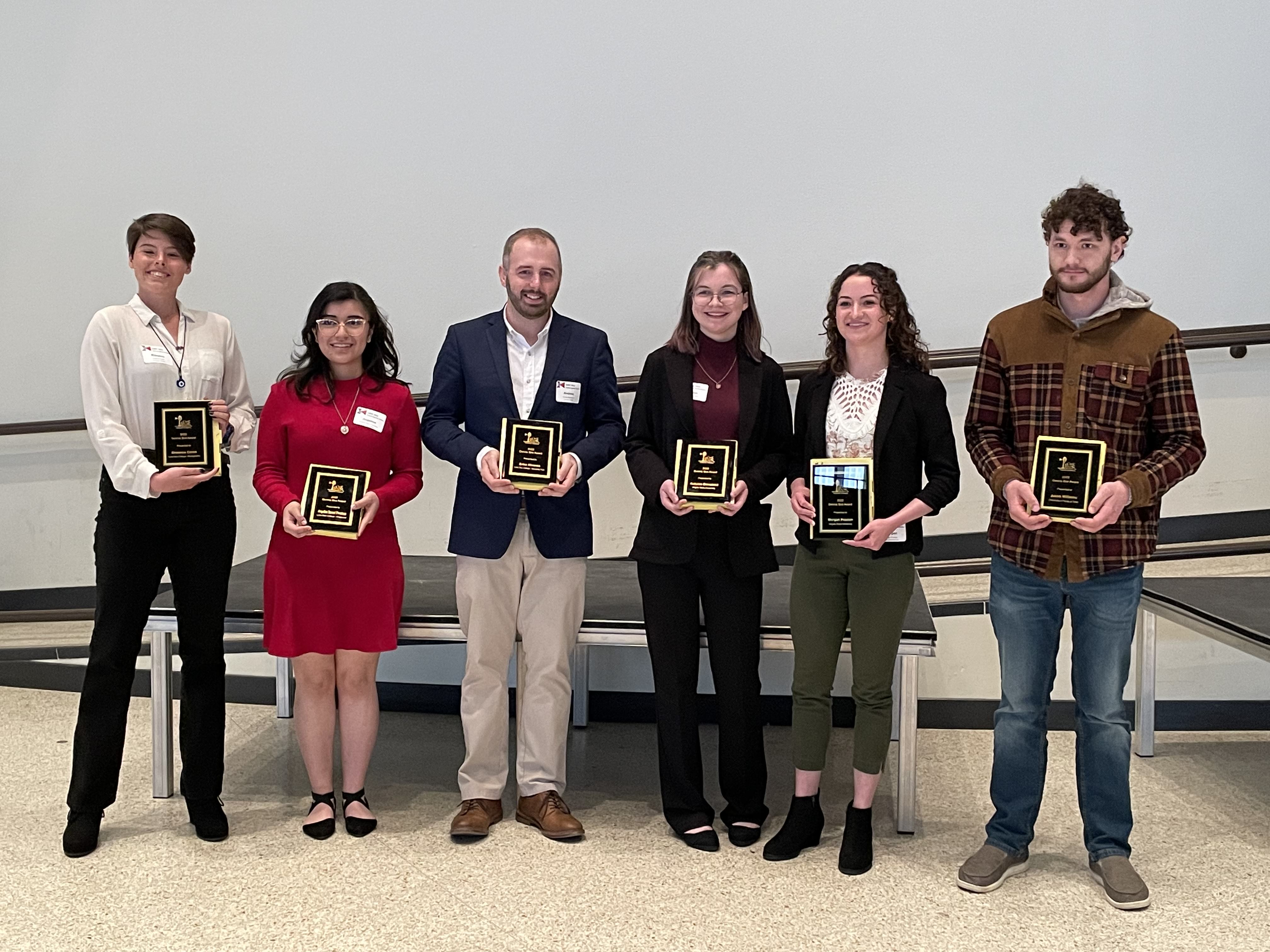 Six individuals standing and each is holding an award plaque
