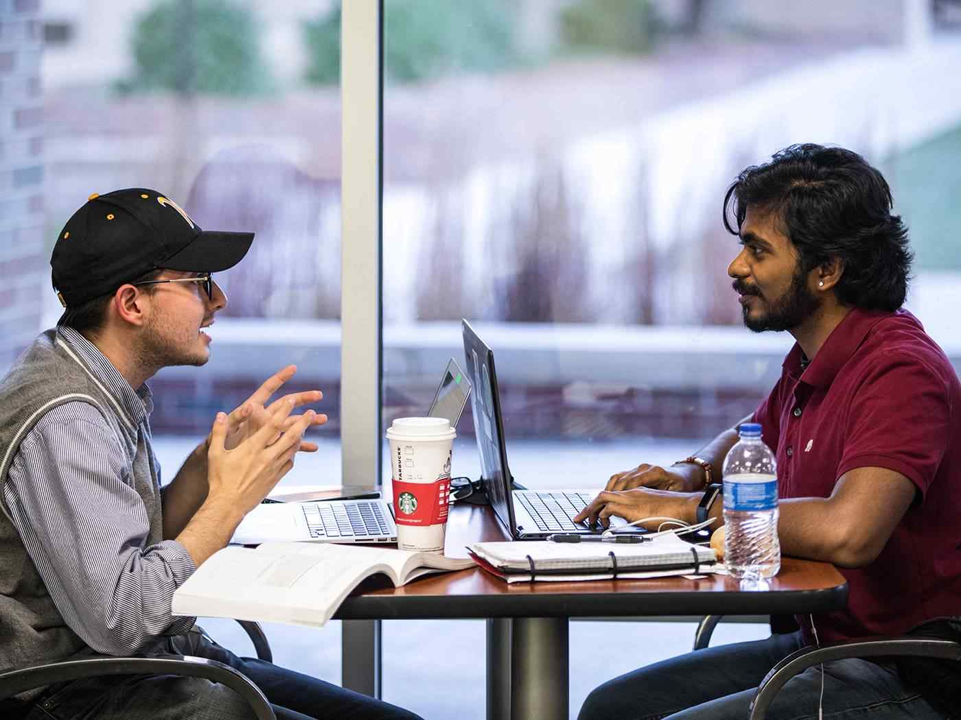 Two communications students sit at opposite ends of a table, both with laptop computers open, engaged in lively discussion. 