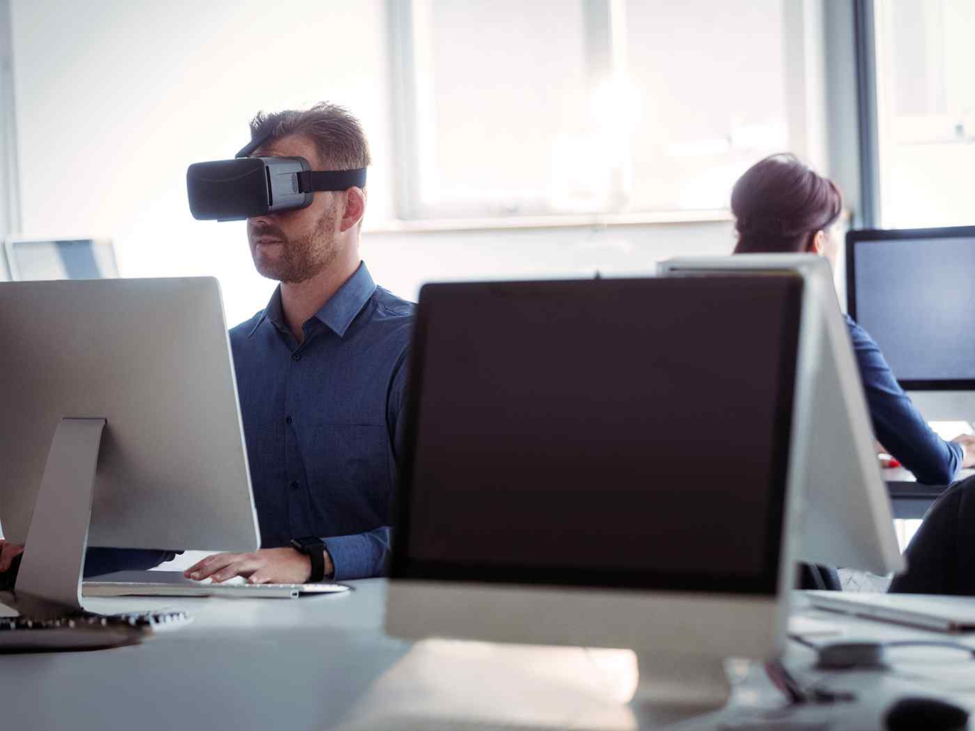 Female and male students seated at computers, wearing virtual reality headsets 