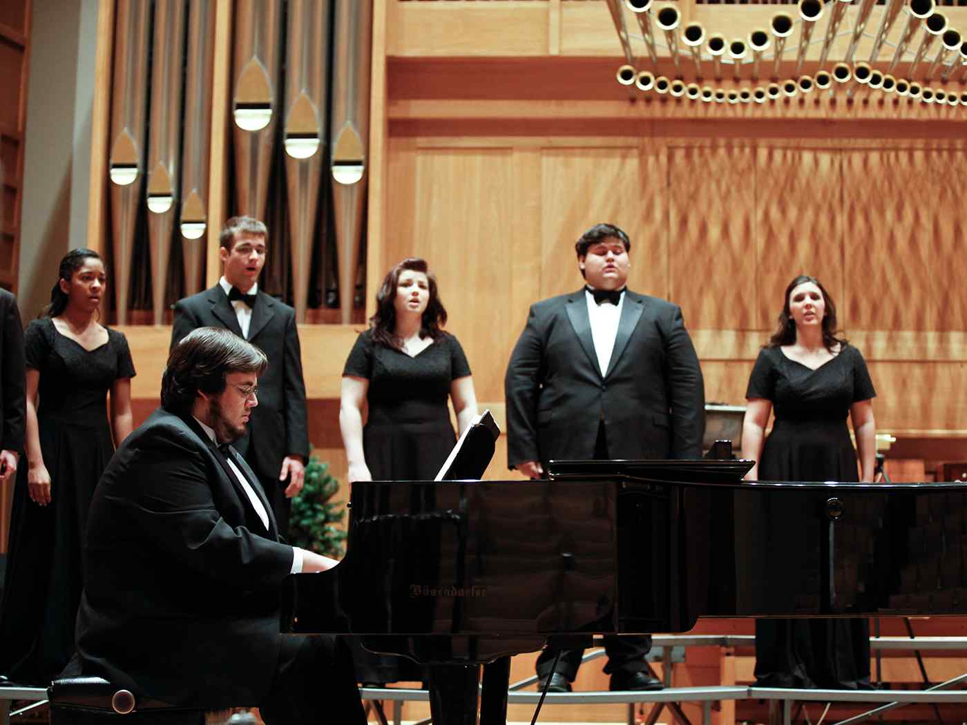 Male student playing the piano while being accompanied by other singing students.