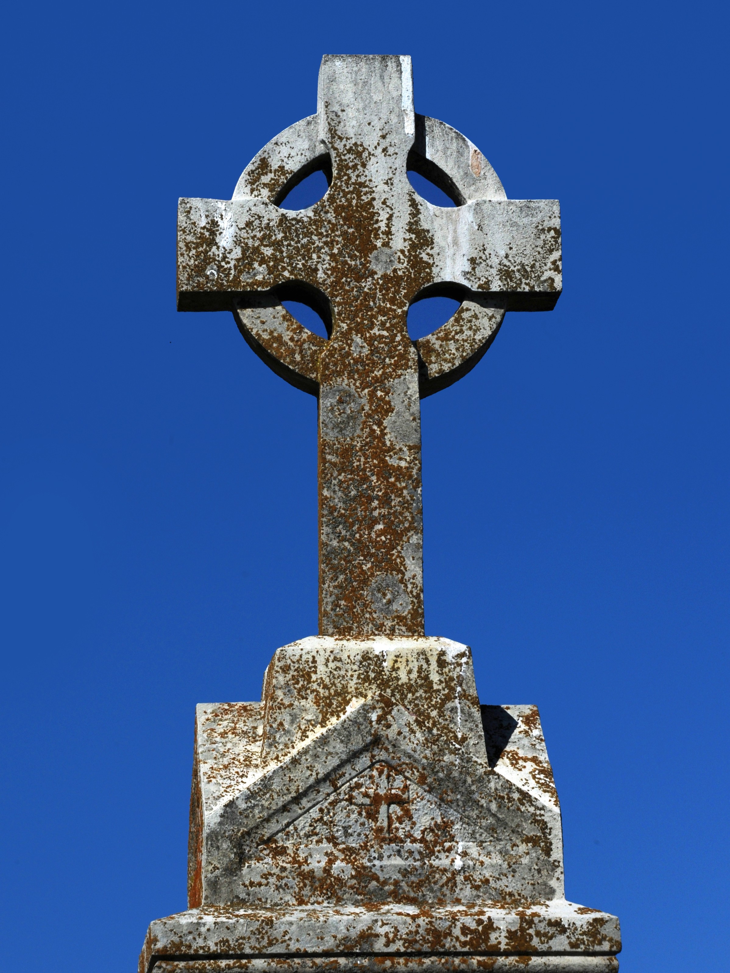A weathered Celtic cross stands against a clear blue sky, its stone covered in patches of brown lichen, evoking a sense of history and serenity.