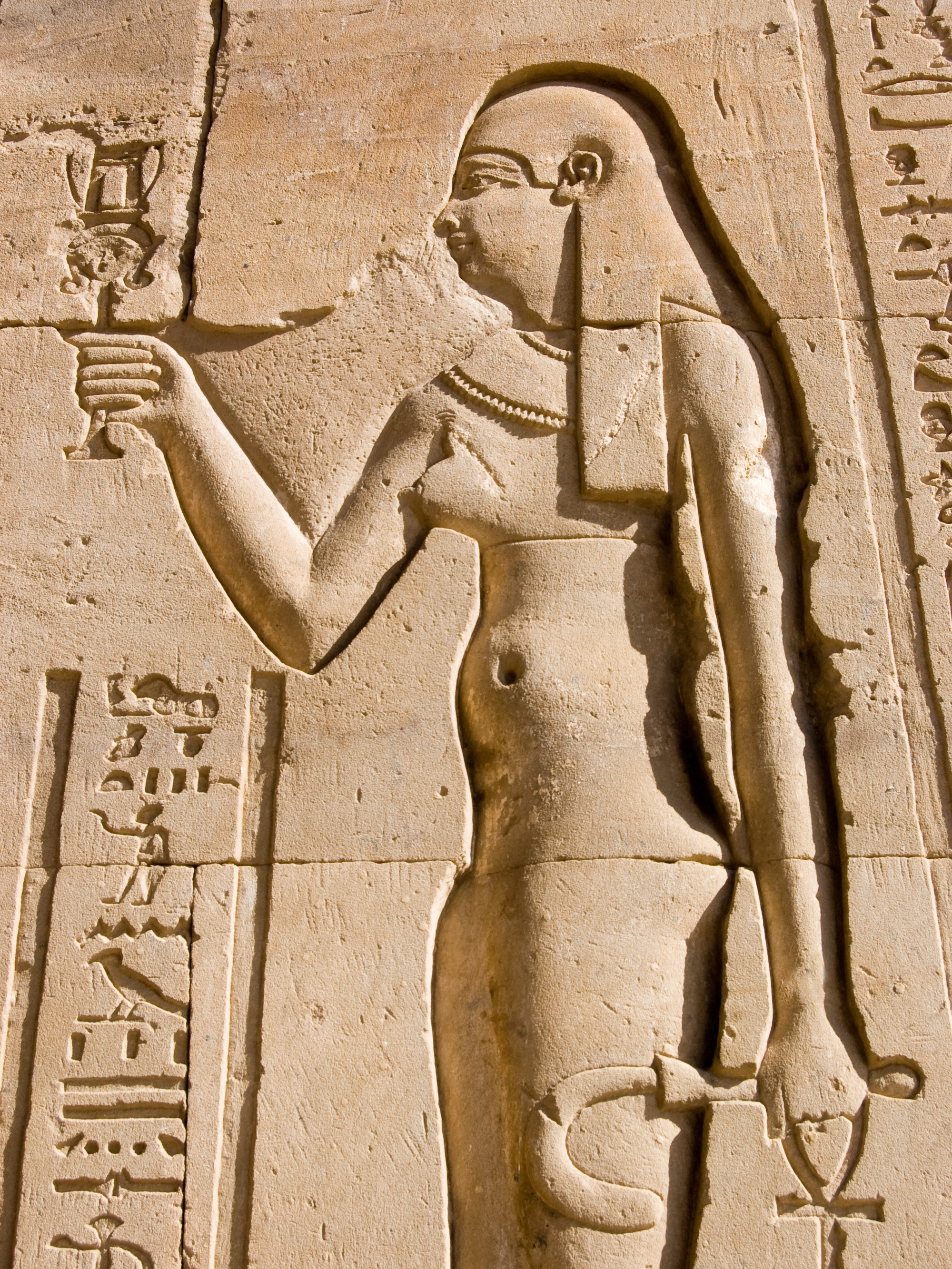 Ancient Egyptian carved relief showing a standing figure with a headdress, holding an ankh and offering chalice. Hieroglyphics are inscribed beside the figure.