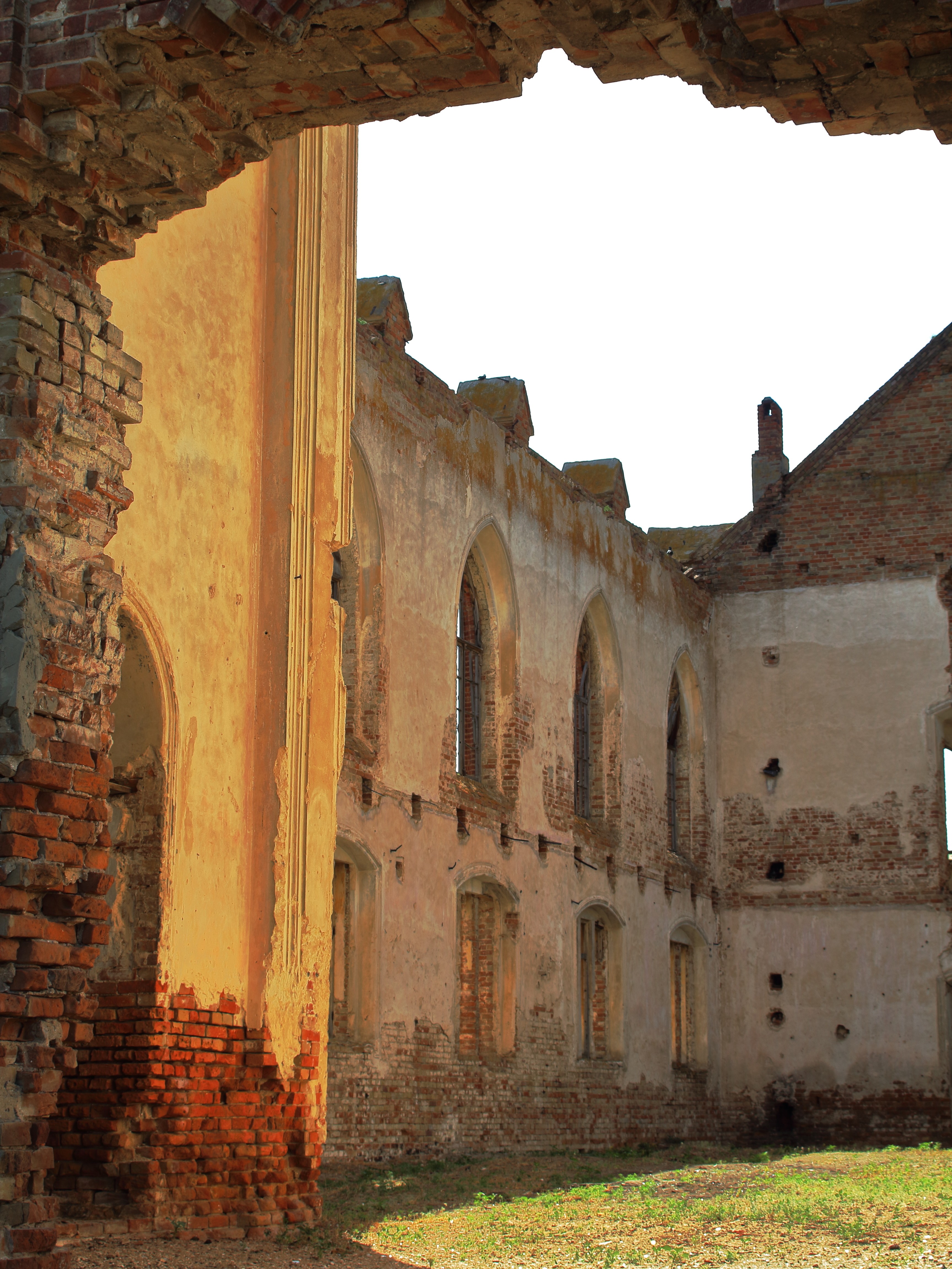 Ruined brick and stone structure with arched windows stands under a bright sky. The sunlight highlights its weathered textures and historical feel.