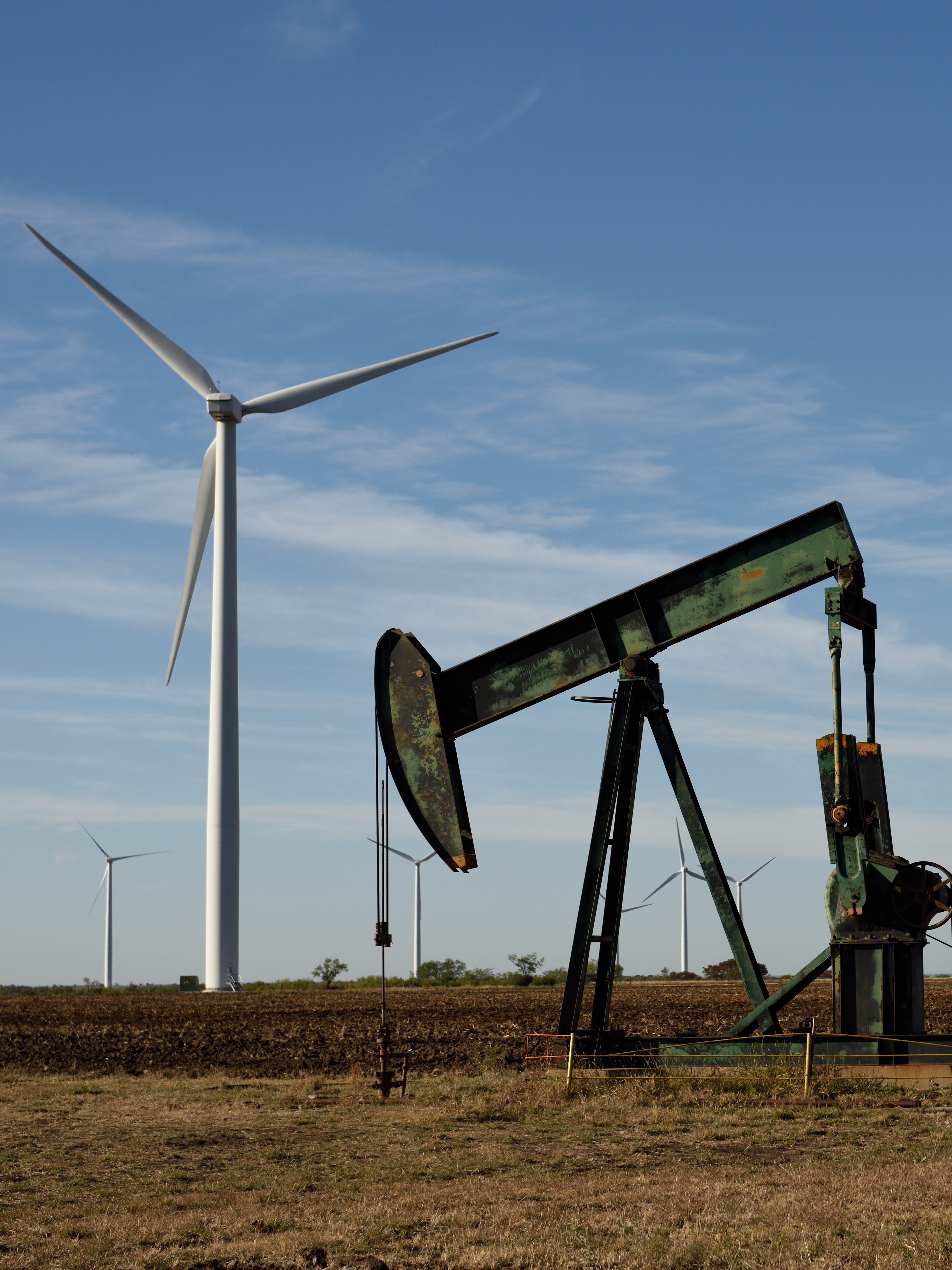 An oil pump jack stands in the foreground of a grassy field, with modern wind turbines in the background under a clear blue sky