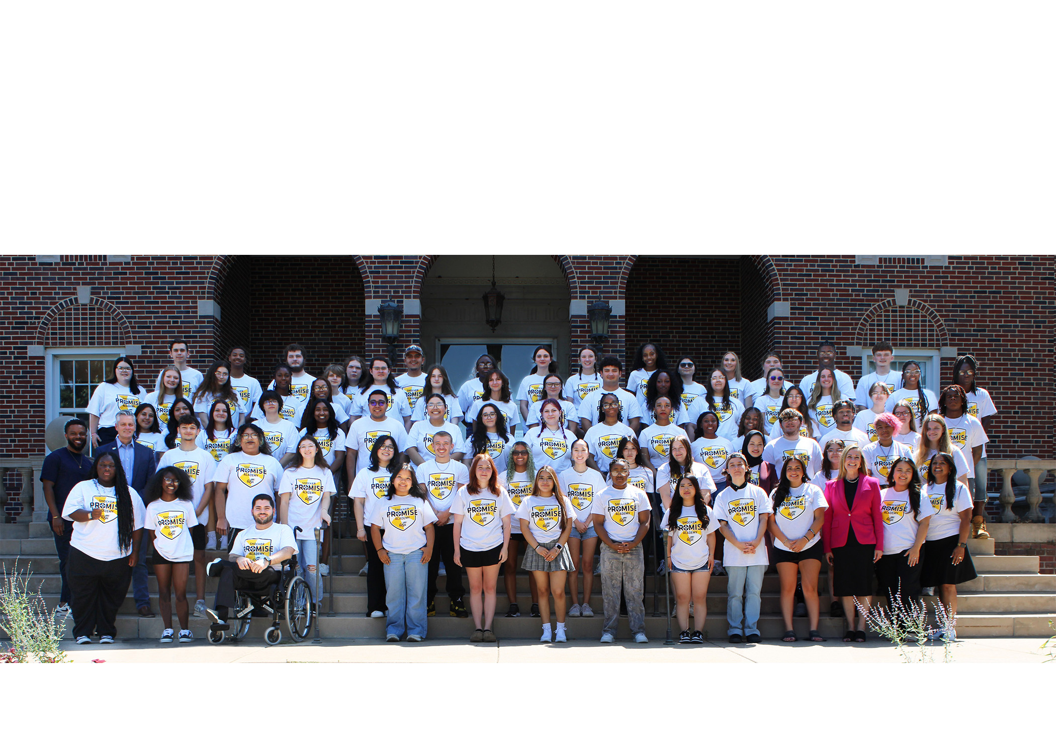 A group photo of the Shocker Promise Academy Scholars posing in front of a WSU building outdoors. All of the students are wearing the same white Shocker Promise T-shirt. Among the students is WSU president Rick Muma and Financial Aid & Scholarships executive director, Sheelu Surender.