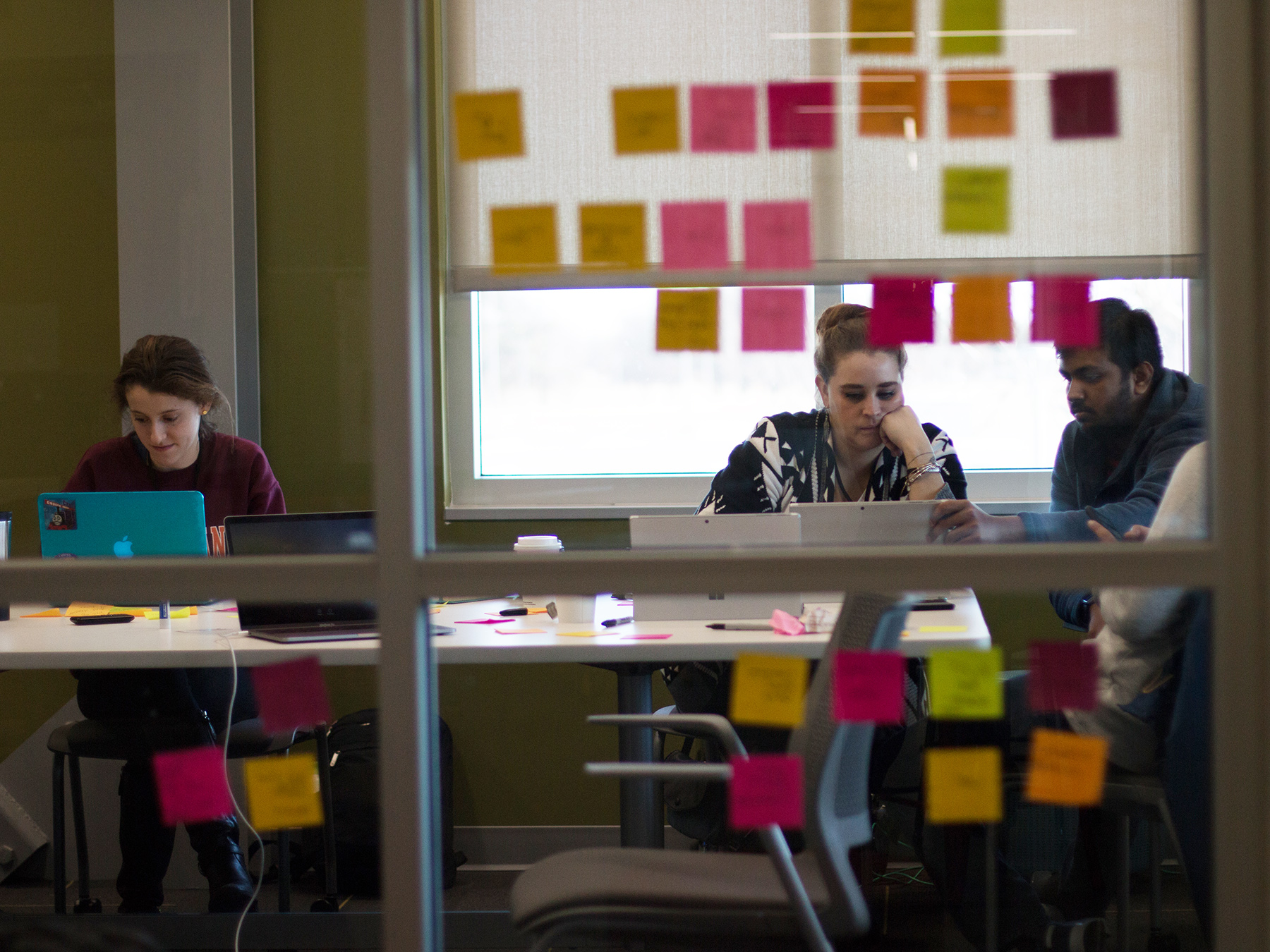 Colorful post-its are visible on a window, in the background students sit at a table working on tablets and laptops