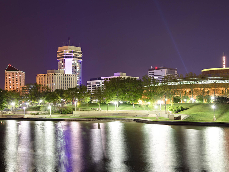 The downtown Wichita skyline, illuminated at night. In the foreground, the Arkansas River is visible.
