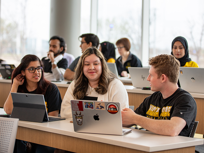 Three students sit with laptops and tablets in a Woolsey Hall classroom