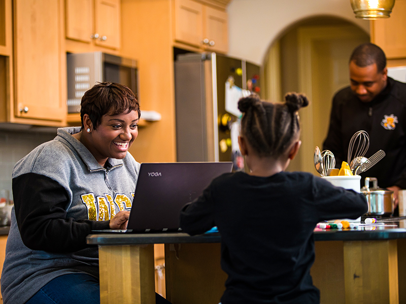 A mom at her kitchen counter studying online while her child plays and dad preps dinner.