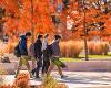 Three students walk in the foreground. Fall foliage visible in the background.