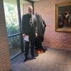 A portrait photo of Dean Rodney Miller standing with the Rigoletto statue just before its reveal. The statue is covered with a black cloth. Dean Miller is wearing a suit and holding some papers.