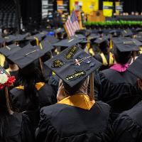 Several graduates facing away from the camera while seated during the commencement ceremony. You can see several decorated caps amongst a sea of standard black caps.