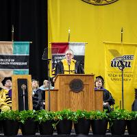 A speaker at the podium for the commencement ceremony. Several other administrators are seated behind the speaker. There are several banners hanging behind the seated people.