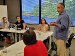 A man stands gesturing as he delivers his pitch to the group.