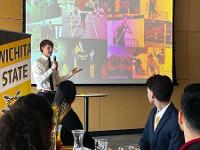 A Gore Scholar stands in front of a projector screen to give a presentation. Visible in the foreground are audience members seated at tables.