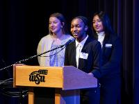 Three Gore Scholars stand at a podium in the CAC Theater as one addresses the audience