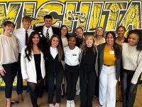 A group of current Gore scholars and Gore alumni pose in front of the yellow Wichita State mural in the Rhatigan Student Center.