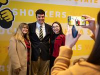 Three students pose against a yellow backdrop while a fourth student takes a photo on their phone.