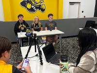 Three students sit at the front of a room during a virtual question and answer session.