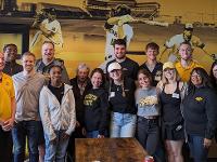 A group of people in Shocker gear pose for a photo in front of a mural depicting Wichita State baseball players at bat and on the field.