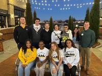 A group of Gore Scholars and Gore Alumni pose for a photo underneath patio string lights outside at Braeburn Square.