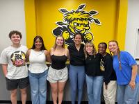 A group of Gore scholars poses in front of a yellow wall decorated with a WuShock.