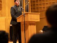 A student speaks at a podium. In the foreground, audience members are visible.