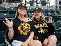 Two students dressed in Shocker gear pose for a photo while seated at a baseball game at Eck Stadium.