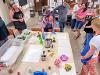 Lori Santos handling pressed floral art with blue latex gloves on a large table filled with various supplies. A group of instructors surround the table.