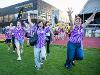 A group of excited students wearing purple tie-dye Fine Arts shirts run and cheer on a grassy field next to a track. Several have face paint and are striking playful poses with arms raised in celebration.