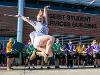 A baton twirler leaps mid-air in front of a cheering crowd and drumline outside the Geist Student Services Building. She is wearing a gray T-shirt and white shorts, holding a baton with a joyful expression.