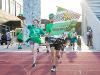 Two students in green College of Engineering shirts run onto a track, one holding a large College of Engineering flag and the other raising a cowboy hat. Other students in green outfits follow behind, with the sun shining brightly.